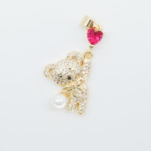 May include: A gold-colored bear pendant with a bow and a pearl. The bear is covered in small, clear crystals. A red heart-shaped gem hangs above the bear. The pendant is on a white background.