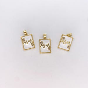 Puede incluir: Tres colgantes dorados con la palabra "mama" en escritura cursiva. Cada colgante tiene un fondo blanco, un borde dorado y pequeños detalles en forma de corazón. Los colgantes son rectangulares con un lazo en la parte superior para una cadena.