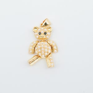 May include: A gold-colored teddy bear pendant with a bail at the top. The bear is adorned with small, round, white pearls. The bear's eyes are black. The pendant is likely for a necklace or bracelet.