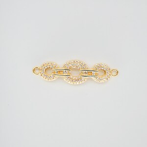 May include: A gold-colored jewelry clasp with three circular links. Each link is encrusted with small, clear crystals. The clasp has small loops on each end for attachment. The background is a solid, light color.