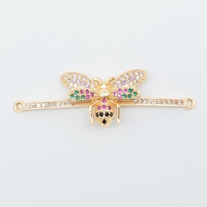 May include: A gold-colored bee-shaped charm with colorful gemstones. The bee has wings with purple, white, and green stones, a pink and black abdomen, and is attached to a bar with clear stones. The charm has small loops on each end.