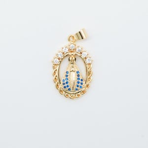 May include: Gold-colored pendant featuring a religious figure with a blue and white design. The pendant is oval-shaped, surrounded by a halo of clear stones and a decorative border. The bail is also gold-colored.