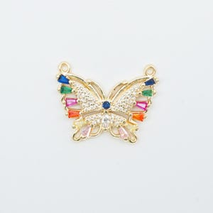 May include: A gold-colored butterfly pendant with colorful, sparkling accents. The butterfly's wings feature a rainbow of colors, including red, orange, yellow, green, and blue. The body is encrusted with clear crystals. Two small loops are at the top for attaching to a chain.