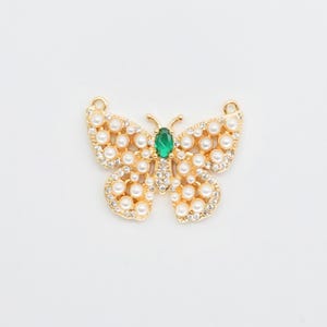 May include: A gold-colored butterfly pendant with a central green gemstone and pearl accents. The butterfly's wings are adorned with small, round pearls and clear crystals, creating a sparkling effect. The pendant has a delicate, ornate design.
