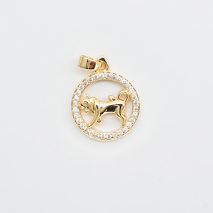 May include: Gold-colored pendant featuring a bull zodiac symbol. The bull is in the center of a circle of small, clear stones. The pendant has a loop at the top for a chain. The background is white.
