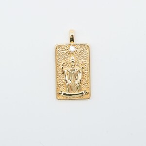 May include: Gold-coloured pendant with a rectangular shape and a detailed relief of a figure in robes. A small, clear stone is set at the top. The pendant has a textured background and a banner at the bottom.