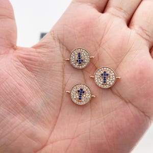 May include: Three gold-colored, round charms with a cross design. The crosses are made of blue gemstones and surrounded by clear gemstones. The charms are small and delicate, suitable for jewelry making.