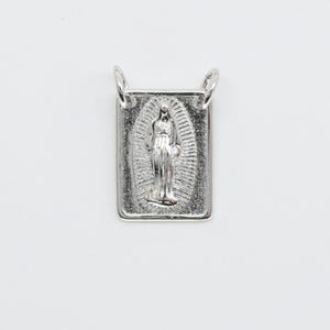 Puede incluir: Un colgante rectangular plateado con una representación detallada de una figura religiosa. El colgante tiene bucles en la parte superior para sujetarlo a una cadena o collar. La figura está rodeada por un diseño ovalado.