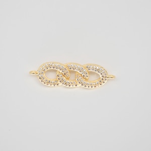 Puede incluir: Un conector de joyería dorado con tres eslabones ovalados entrelazados. Cada eslabón está incrustado con pequeños cristales transparentes, creando un efecto brillante. El conector tiene pequeños bucles en cada extremo para la fijación.