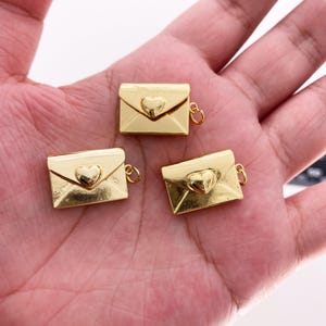 以下が含まれることがあります： ハートのアクセントが付いた、3つの金色の封筒チャーム。各チャームには、ジュエリーやその他の工芸品に取り付けるための小さなループが付いています。封筒は長方形で、中央にハートが付いています。