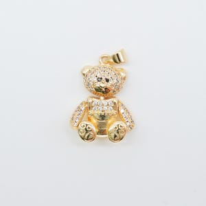 May include: A gold-colored teddy bear pendant with sparkling clear crystals. The bear has a small loop at the top for a chain. The bear's head and arms are covered in crystals, and the body has a smooth, polished finish.