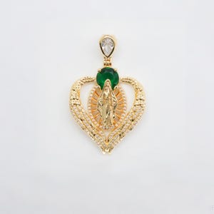 May include: Gold-toned pendant featuring a heart-shaped design with a central figure and a green gemstone. The pendant has a teardrop-shaped clear stone at the top and is adorned with small, sparkling accents. A religious-themed jewelry piece.