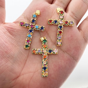Könnte beinhalten: Drei goldfarbene Kreuzanhänger, jeder mit einer lebendigen Anordnung farbiger Edelsteine verziert. Die Edelsteine sind in einem Muster angeordnet, das einen Regenbogeneffekt erzeugt. Die Kreuze werden auf hellem Hintergrund gezeigt.