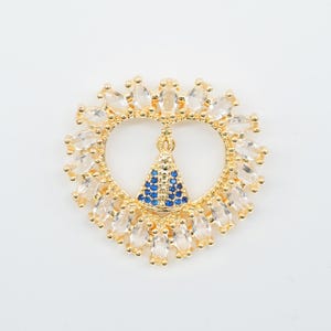 May include: Gold-toned pendant with a heart-shaped design, featuring clear and blue gemstones. The pendant has a central figure with blue stones and is surrounded by clear, oval-shaped stones. The overall design is ornate and detailed.