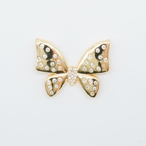 May include: A gold-colored bow-shaped brooch with a central cluster of small, clear crystals. The bow's wings are adorned with evenly spaced, smaller clear crystals, creating a sparkling effect. The brooch is set against a white background.