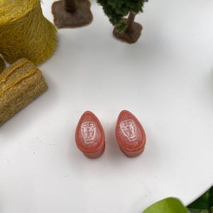 May include: A pair of pink teardrop-shaped ear gauges with a white drum design.