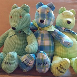 May include: Three stuffed teddy bears in shades of green and blue plaid fabric. Each bear has a bow tie or collar and the words "Your Angel" and "PAPA" embroidered on their feet. The bears are sitting on a neutral-colored couch.