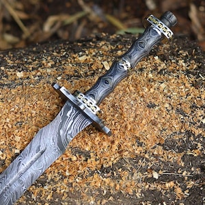 May include: A detailed close-up of a decorative dagger with a patterned blade and handle. The handle is dark with silver accents and a skull design. The blade has a unique pattern. The dagger is resting on a wood surface.