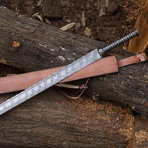 May include: A Damascus steel sword with a patterned blade and a textured handle lies on a brown leather sheath. The sword is displayed on weathered logs, highlighting its craftsmanship.