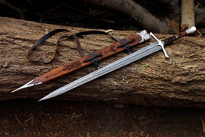 May include: A detailed image of a sword and scabbard resting on a log. The sword has a silver blade with a patterned design, a silver crossguard, and a dark wooden handle. The scabbard is made of wood with leather straps and decorative carvings.
