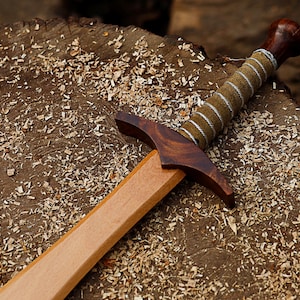 May include: A wooden toy sword with a light brown blade and a dark brown hilt. The handle is wrapped in tan fabric with silver-colored wire. The sword rests on a wood surface covered in wood shavings.