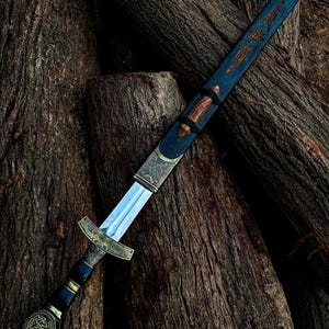 May include: An ornamental sword with a silver blade and an elaborate gold and black handle. The blue scabbard has gold detailing and a wooden grip. The sword is displayed on a pile of weathered logs, highlighting its craftsmanship.