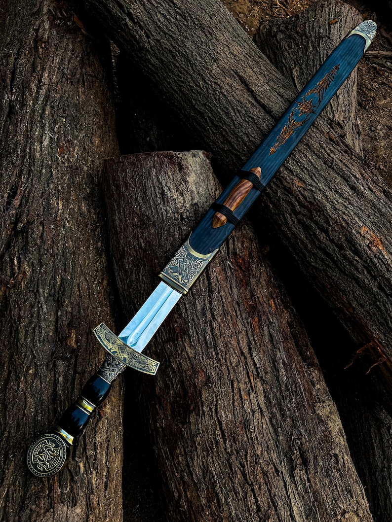 May include: An ornamental sword with a silver blade and an elaborate gold and black handle. The blue scabbard has gold detailing and a wooden grip. The sword is displayed on a pile of weathered logs, highlighting its craftsmanship.