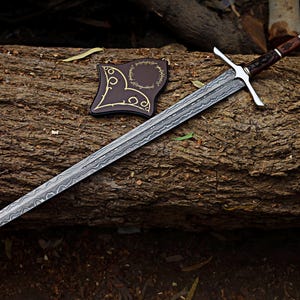 May include: A long, silver sword with a patterned blade and a cross-shaped hilt rests on a log. The handle is brown with silver accents. A brown leather sheath with gold detailing is next to the sword. The items are set against a natural wood background.