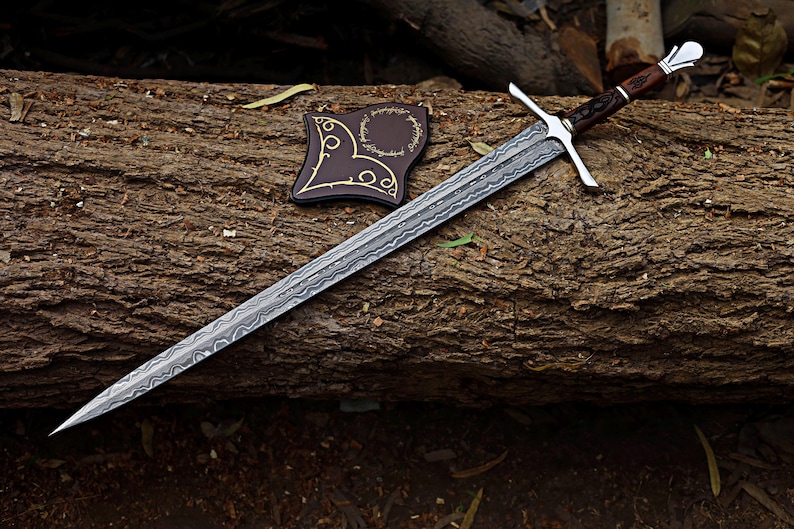 May include: A long, silver sword with a patterned blade and a cross-shaped hilt rests on a log. The handle is brown with silver accents. A brown leather sheath with gold detailing is next to the sword. The items are set against a natural wood background.