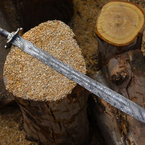 May include: An intricately designed sword with a patterned blade, resting on a wooden surface. The handle is dark with gold detailing. The image highlights the sword's craftsmanship and the materials used.