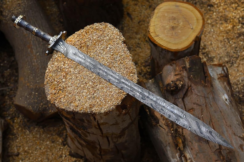 May include: An intricately designed sword with a patterned blade, resting on a wooden surface. The handle is dark with gold detailing. The image highlights the sword's craftsmanship and the materials used.