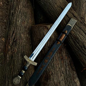 May include: A silver sword with an ornate gold hilt and a black and gold scabbard rests on a bed of brown logs. The sword's blade is polished, reflecting light. The scabbard features detailed carvings and gold accents, showcasing a historical or fantasy theme.