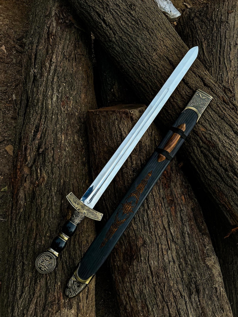 May include: A silver sword with an ornate gold hilt and a black and gold scabbard rests on a bed of brown logs. The sword's blade is polished, reflecting light. The scabbard features detailed carvings and gold accents, showcasing a historical or fantasy theme.