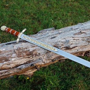 Peut inclure: Une épée à lame argentée avec un manche en cuir marron et des détails dorés repose sur une bûche patinée. La lame présente un texte gravé et une étoile décorative. L'épée est un objet historique ou fantastique.
