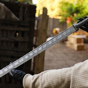 May include: A patterned metal sword with a textured handle. The blade has a unique design and is held by a person wearing black gloves. The sword is displayed outdoors. The blade's length is approximately 60 cm.