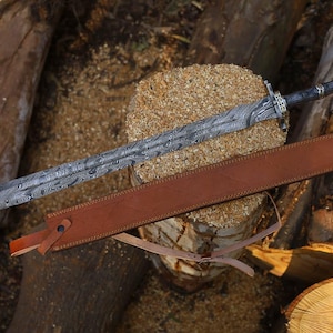 May include: An ornate sword with a patterned blade and a dark handle, lying on a wooden surface. A brown leather sheath accompanies the sword. The blade's length is approximately 76 cm.