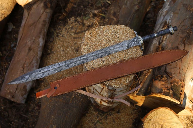 May include: An ornate sword with a patterned blade and a dark handle, lying on a wooden surface. A brown leather sheath accompanies the sword. The blade's length is approximately 76 cm.