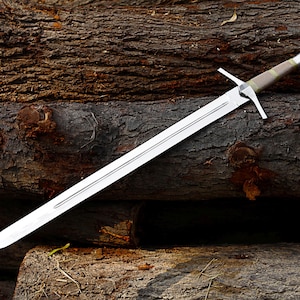 May include: A long, silver sword with a cross-shaped hilt and a brown and green handle rests on a pile of rough, brown logs. The blade is sharp and the hilt is silver. The sword is a medieval weapon.