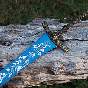 May include: A decorative sword with a blue blade featuring a white floral design. The hilt and crossguard have a gold and bronze woven pattern. The sword rests on a weathered wooden log, with green grass in the background.
