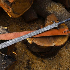 May include: A patterned steel sword with a dark handle and a leather sheath. The sword is displayed on a wooden surface, highlighting its detailed design and craftsmanship. The blade features a distinctive pattern.
