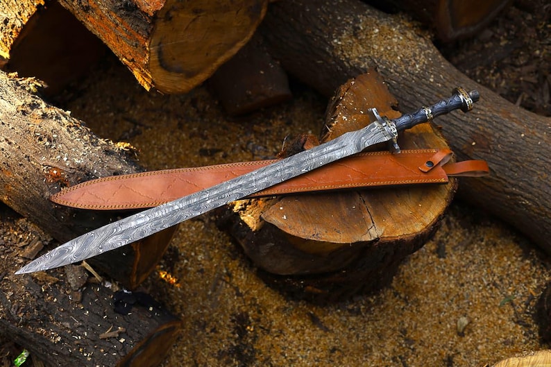 May include: A patterned steel sword with a dark handle and a leather sheath. The sword is displayed on a wooden surface, highlighting its detailed design and craftsmanship. The blade features a distinctive pattern.
