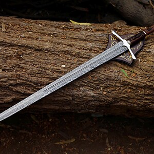 May include: A long, silver sword with a patterned blade rests on a wooden log. The sword features a brown handle, silver crossguard, and a decorative leather sheath. The blade has a detailed, etched design.