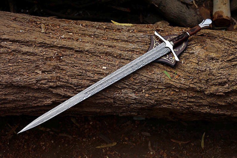 May include: A long, silver sword with a patterned blade rests on a wooden log. The sword features a brown handle, silver crossguard, and a decorative leather sheath. The blade has a detailed, etched design.