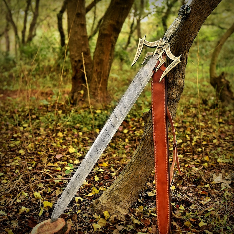 May include: A silver sword with a gold-colored hilt and a leather strap. The sword is leaning against a tree trunk in a forest setting.