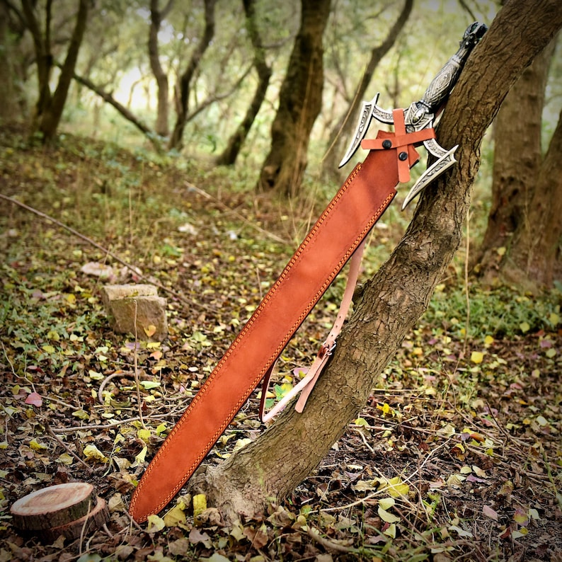 May include: A brown leather sword scabbard with a silver sword with a decorative handle. The scabbard has a strap and is resting against a tree trunk in a forest setting.