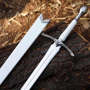 May include: A close-up of a sword with a white handle and a silver blade, placed on a dark wooden surface. The sword has a white sheath. The handle is wrapped in white material, and the crossguard is silver.