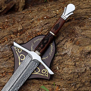 May include: A detailed sword with a patterned blade and a wooden handle. The handle has intricate carvings and is set on a brown plaque with gold accents. The sword is displayed on a textured, brown surface.