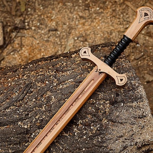 May include: A wooden sword with a light brown blade and handle, featuring a black grip and decorative carvings. The sword rests on a textured, brown log, with wood shavings in the background. The blade has engraved text.