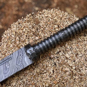 May include: Close-up of a Damascus steel knife with a textured handle. The blade displays a unique wood grain pattern. The handle is dark grey with a spiral design. The knife rests on a bed of wood shavings.
