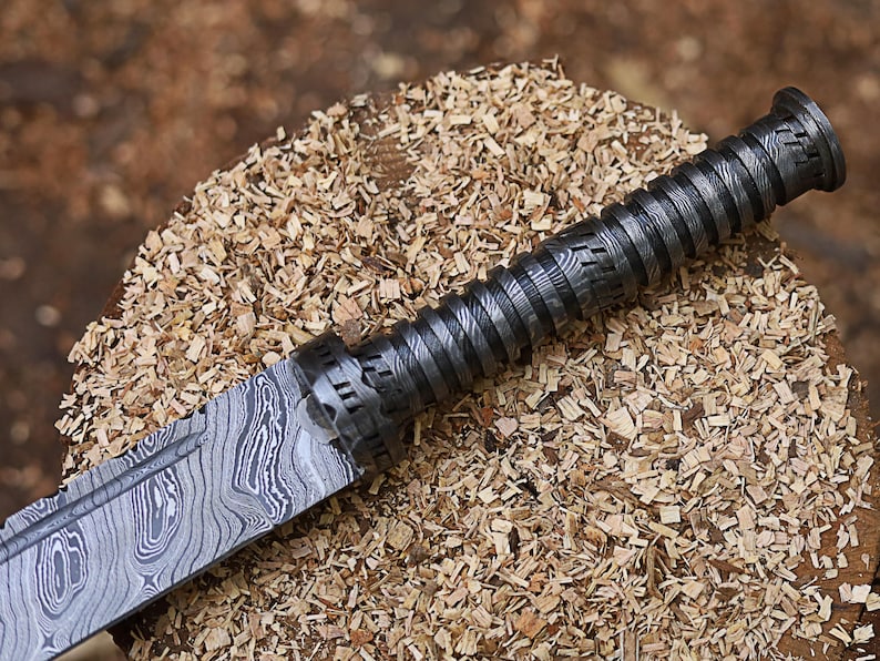 May include: Close-up of a Damascus steel knife with a textured handle. The blade displays a unique wood grain pattern. The handle is dark grey with a spiral design. The knife rests on a bed of wood shavings.